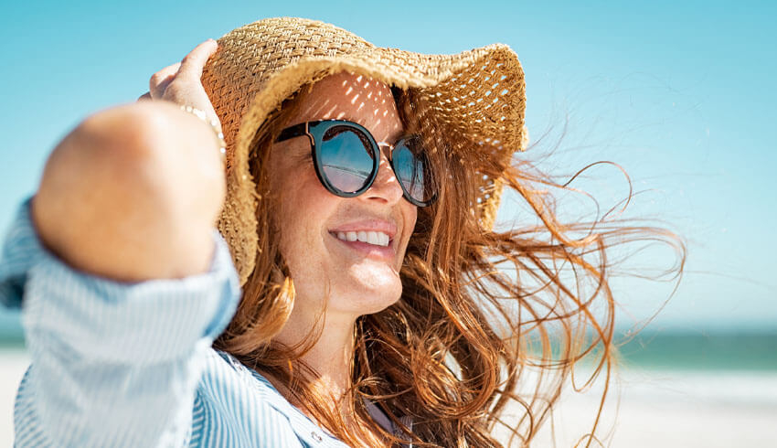 A woman with red hair and fair skin wears sunglasses and a sun hat on the beach on a sunny day.
